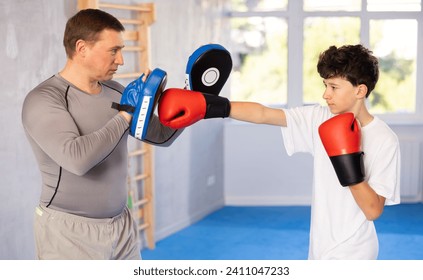 Sportive teenage boy training boxing kicks on punch mitts held by instructor in sports hall - Powered by Shutterstock