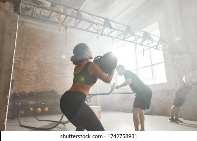 Sportive Woman Lifting A Heavy Sandbag While Having Workout At Industrial Gym. Group Training, Teamwork Concept. Selective Focus