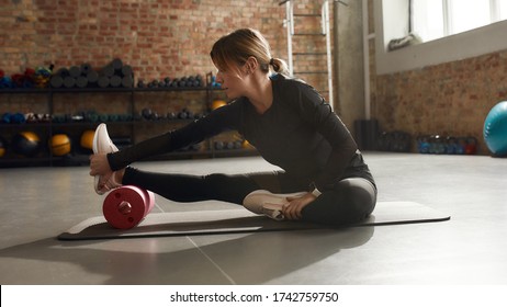 Sportive woman sitting on a yoga mat, using foam roller while having workout at industrial gym. Healthy lifestyle concept. Horizontal shot. Side view - Powered by Shutterstock