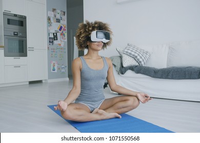 Sportive young woman sitting on blue mat at home relaxing and meditating in glasses of virtual reality looking content. - Powered by Shutterstock