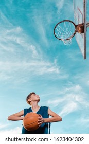 Sports and basketball. A young teen in a black tracksuit playing basketball on the school Playground. Cloudy blue sky with basket on the background. View from the bottom. Vertical. Copy space