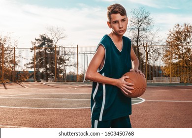 Sports and basketball. A young teenager in a blue tracksuit poses with a basketball in his hands. In the background, a basketball court. Horizontal. Close up