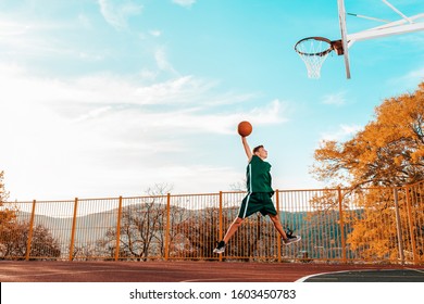 Sports and basketball. A young teenager in a green tracksuit jumps and throws a ball into the basket. Blue sky and court in the background. Copy space - Powered by Shutterstock