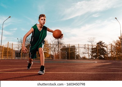 Sports and basketball. A young teenager in a green tracksuit plays basketball, leads the ball before throwing. Blue sky in the background and a sports field in the background. Horizontal. Copy space