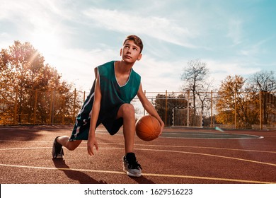 Sports and basketball. A young teenager in a green tracksuit playing basketball, leading the ball. Blue sky in the background and a sports ground in the background. Copy space