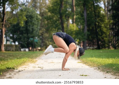 Sports blonde yoga practitioner, performs Bakasana exercise, crane pose, handstand, practices in shorts and tank top on park footpath, full-length side view - Powered by Shutterstock