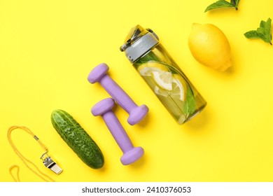Sports bottle of lemonade with cucumber and dumbbells on yellow background
