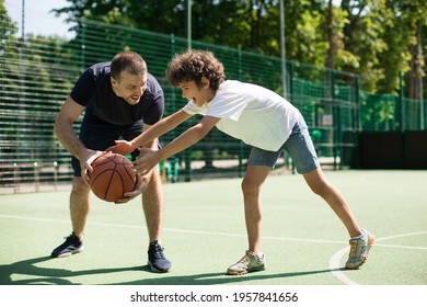 Sports Education. Full body length portrait in action of adult man and young boy playing basketball on a court, coach leading the ball and dribbling, teaching little player, spending time outdoors - Powered by Shutterstock