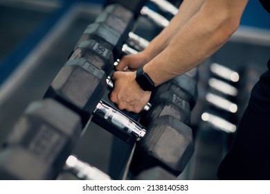 Sports equipment concept. Unrecognizable muscular man taking dumbbells from the rack in gym, cropped shot of male athlete grabbing barbells, preparing for workout with light weights, closeup