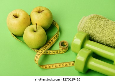 Sports And Healthy Regime Equipment, Copy Space. Barbells Near Juicy Green Apples. Athletics And Weight Loss Concept. Dumbbells In Bright Green Color, Measure Tape, Towel And Fruit On Green Background