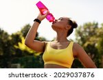 Sports And Hydration Concept. Portrait of fit black woman drinking fresh water from bottle, wearing yellow sportswear top bra, exercising oudoors taking break. Street Workout, Outside Training