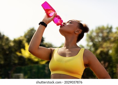 Sports And Hydration Concept. Portrait of fit black woman drinking fresh water from bottle, wearing yellow sportswear top bra, exercising oudoors taking break. Street Workout, Outside Training