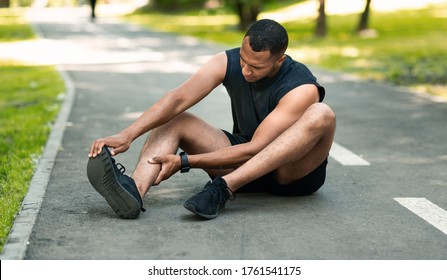 Sports injury. African American runner sitting on jogging track and feeling pain in his ankle, panorama