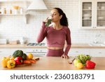 Sports Nutrition. Slim Young Woman In Fitness Fitwear Drinking Green Smoothie, Enjoying Detox Drink Posing With Glass At Modern Kitchen At Home, Standing Near Table With Fruits And Vegetables