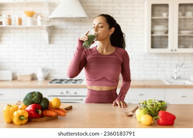 Sports Nutrition. Slim Young Woman In Fitness Fitwear Drinking Green Smoothie, Enjoying Detox Drink Posing With Glass At Modern Kitchen At Home, Standing Near Table With Fruits And Vegetables - Powered by Shutterstock