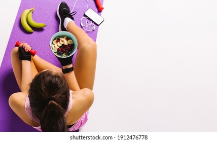 Sports and nutrition. Top photo of a girl with dumbbells and muesli in hand while sitting on the mat. White background and blank advertising space.