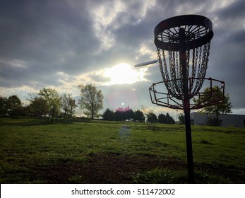 Sports Photo of a Disc Golf Disc Flying into a Chain Link Basket - in Silhouette with Rays of Sun Shining Through Heavy Clouds on a Weekend Morning
