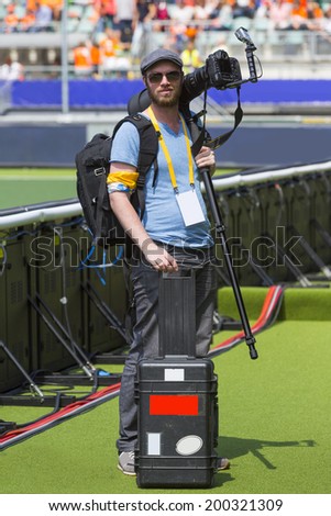 A sports photographer posing along a sportsfield with his camera on his shoulder with a monopod mounded on it, in his other hand he hold a case for the other lenses. 