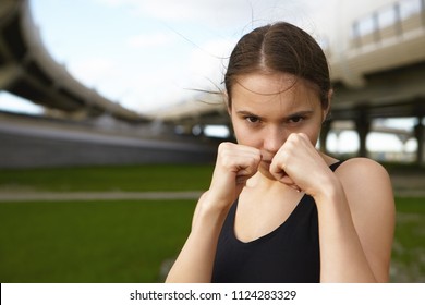 Sports, strength, self defense and martial arts concept. Close up shot of strong serious sporty girl boxing outdoors, holding clenched fists at her face, going to punch you, having focused look