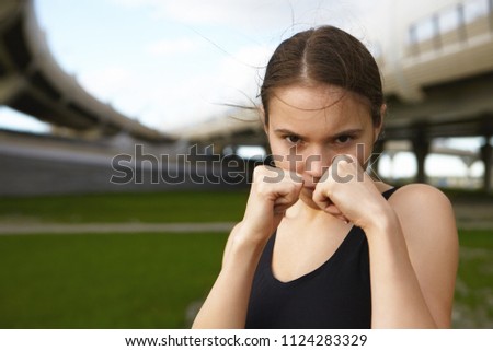 Sports, strength, self defense and martial arts concept. Close up shot of strong serious sporty girl boxing outdoors, holding clenched fists at her face, going to punch you, having focused look