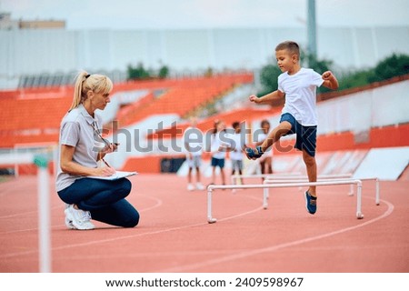 Sports teacher using stopwatch while boy is jumping over hurdles on running track. Focus is on kid. 