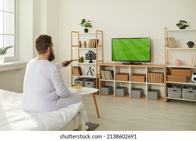 Sports, Television, Entertainment. Relaxed Lazy Male Bearded Fat Football Fan In Pajamas Watching Football Match On TV. Chubby Man Sits On Bed At Home And Turns On Football On TV Using Remote Control.