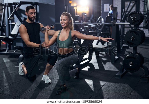 Sports trainer dressed in\
black raising an athlete arm with dumbbell a bit for easier\
exercise performance