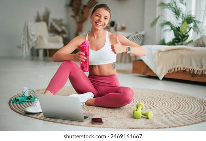 Sports trainer or teacher of Pilates online. Happy blond female in sportswear sitting on the floor with laptop after exercises and showing her thumb up on camera with a smile. - Powered by Shutterstock