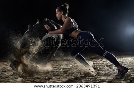 Sports woman turns over tire wheel in gym, spraying sand. Concept workout, zeal for victory, overcoming yourself. Crossfit woman exercising with big tire.