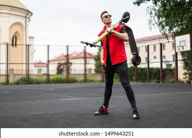 Sports Young Hipster Man In Trendy Denim Clothes In Sneakers Stands With A Scooter On Shoulder On A Basketball Court In The City. Guy In Sunglasses Is Resting After Riding An Electric Scooter Outdoors