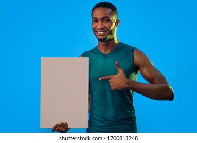 Sportsman Pointing Finger At Blank White Poster Advertising Gym Over Turquoise Blue Background In Studio. Mockup