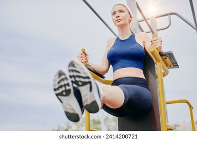 Sportsman in a simple sportswear raising her feet while exercising on the bars, stretching outdoor in summertime, preparing for workout, core muscles strength. Side view - Powered by Shutterstock