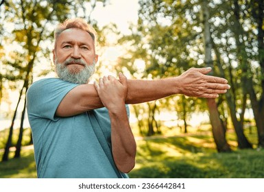 Sportsman working out on nature. Portrait of charismatic athletic man of mature age doing arms exercises for warming up outdoors. Healthy man dressed in blue shirt confidently looking at camera. - Powered by Shutterstock