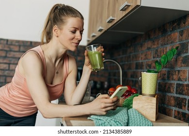 Sportswoman drinking vegan smoothie made of green vegetables and celery in the kitchen and talking on the phone - Shutterstock ID 2134385889