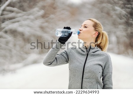 Sportswoman drinking water and taking a break from exercising while standing in woods at snowy winter day. Healthy life, winter fitness, refreshment
