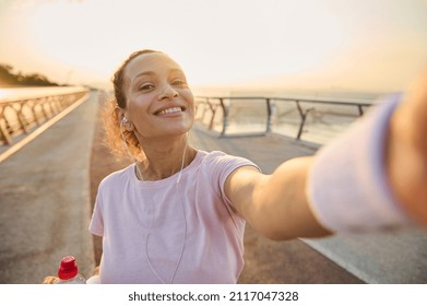 Sportswoman, female athlete in pink t-shirt and wristbands smiles holding a mobile phone in her outstretched hands while making self-portrait during morning jog and workout on the bridge at sunrise - Powered by Shutterstock