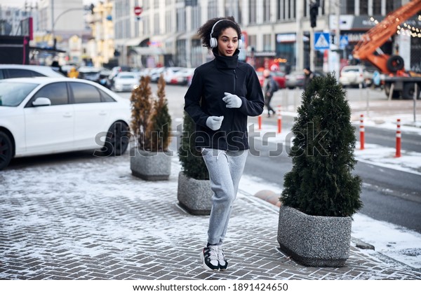 Sportswoman with headphones jogging near the\
bushes on a busy street filled with\
snow