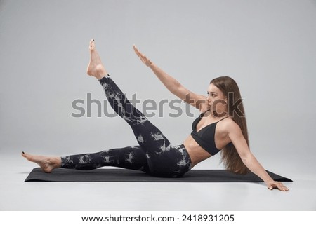 Sportswoman with long fair hair doing crunches in studio. Side view of young Caucasian female raising arm and leg, while having abs workout, isolated on grey background. Concept of sporty lifestyle. 
