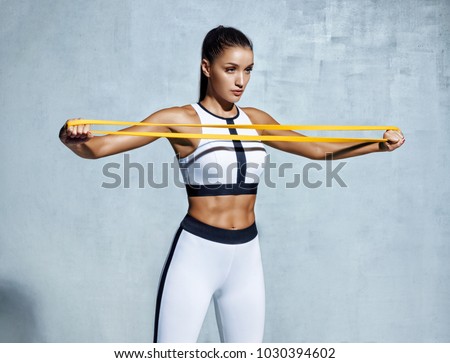 Sportswoman performs exercises for the muscles of the chest. Photo of young woman workout with resistance band on grey background. Strength and motivation.