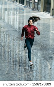 Sportswoman Running Wearing A Red Windbreaker, Black Leggings And White Trainers Under The Rain In The City