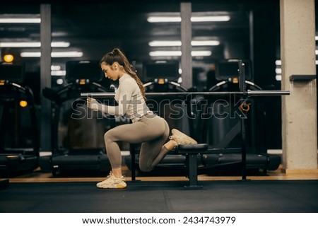 A sportswoman in shape is doing split squats on a bench in a gym.