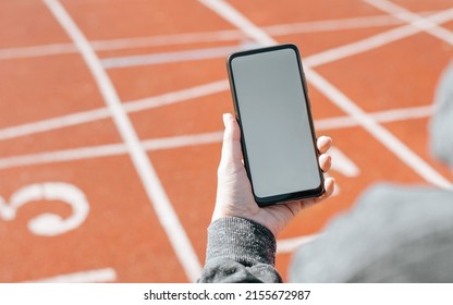 Sportswoman Using Mobile Phone Mockup In Stadium Outdoors. Close-up Of Woman's Hand Holding Smartphone With Blank White Screen With Empty Advertising Space. Sports And Technology Concept.