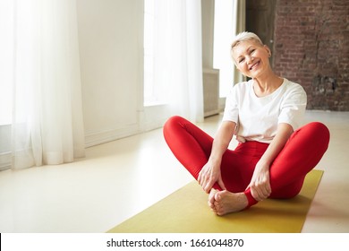 Sporty 50 year old gray haired woman sitting barefooted on yoga mat indoors doing bound ngle pose which helping to relieve symptoms of menopause. Aging, maturity, wellness and health concept
