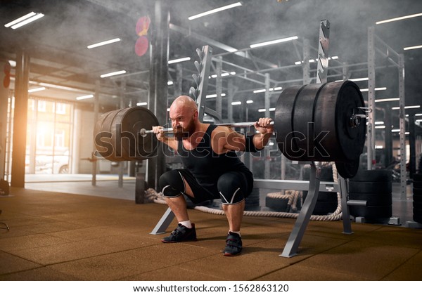 Sporty active man with\
strong arms and severe face, trying to stand with heavy barbell on\
hands, strong powerlifter training in gym, white smoke in the air,\
side indoor shot
