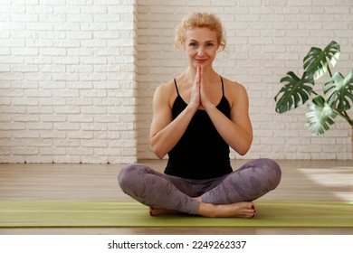 Sporty adult woman sitting in lotus pose with her palms pressed together in front of the chest. Fit middle aged yogini practicing yoga and meditation at home. Background, copy space, close up. - Powered by Shutterstock