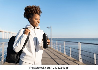 Sporty African American Female With Backpack And Water Bottle Walking Outdoors, Smiling Young Black Woman Ready For Training Outside, Standing On Wooden Pier Near Sea And Looking Aside At Copy Space - Powered by Shutterstock