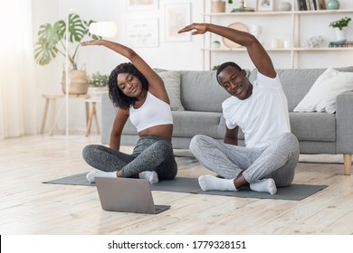 Sporty African Couple Stretching At Home Together, Watching Online Lesson On Laptop