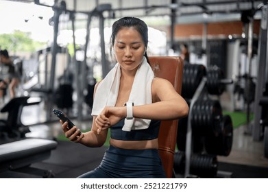 A sporty Asian woman in athletic wear is tracking her burnt calories, checking her workout results on her smartwatch while resting on a bench press at the gym. gadget and sport lifestyle concepts - Powered by Shutterstock