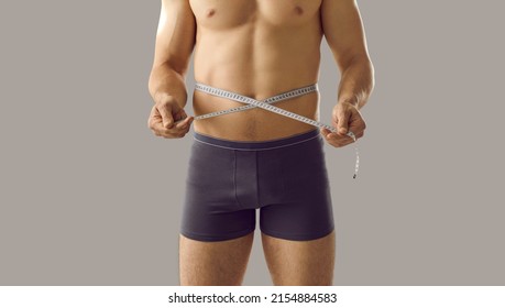 Sporty attractive man in underwear with fit ideal body and beautiful torso standing isolated on grey background holding tape measure on slim waistline to show his workout result. Weight loss concept