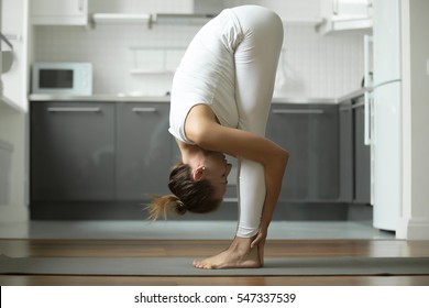 Sporty attractive woman practicing yoga, standing in head to knees exercise, uttanasana pose, working out, wearing white sportswear, indoor full length, home interior background